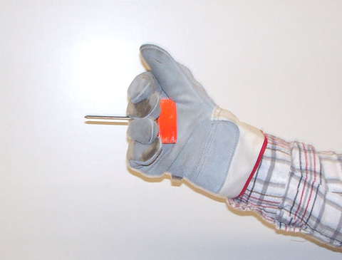 person holding a orange block of wood with a nail; nail is between the 3rd and 4th finger with the point sticking out and the nail head against the block