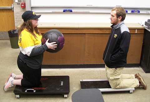 woman kneeling on a cart holding a medicine ball, man kneeling on a cart across from her 